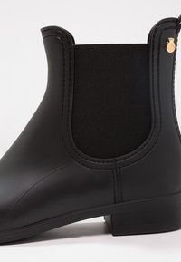 Bottine en caoutchouc noir avec panneau latéral élastique, petit talon bloc et embellissement doré sur le côté extérieur. Texture lisse, design épuré.