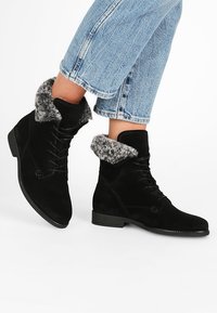 Bottines en daim noir avec un revers en fausse fourrure, un design à lacets, une semelle plate et une finition texturée, portées avec un jean bleu clair court.