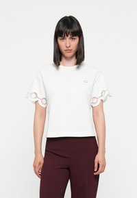 White short-sleeve top with scalloped embroidered accents at sleeves, round neckline, and a small gold logo on the chest.