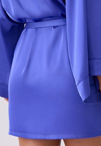 Krótka niebieska satynowa sukienka z paskiem w talii, z długimi rękawami i gładką fakturą, podkreślająca nowoczesny i dopasowany krój.