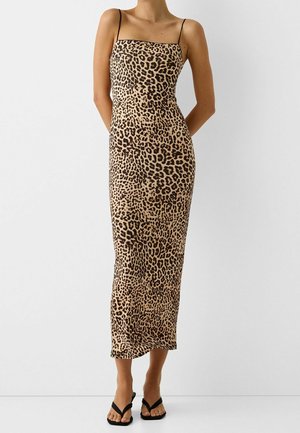 Femme portant une robe midi sans manches à imprimé léopard avec fines bretelles et des sandales noires à bouts ouverts, debout devant un fond blanc uni.
