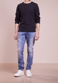 Black long-sleeve shirt, blue distressed jeans, white sneakers. The shirt has a round neckline; jeans are tapered with visible wear.