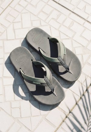 Par de chanclas Havaianas grises con suelas texturizadas descansando sobre un suelo blanco de baldosas con patrón y la sombra de una planta cercana.
