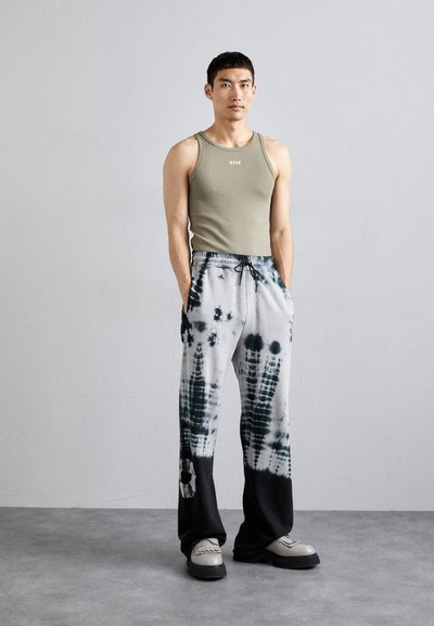 Pantaloni gri și negri, cu efect tie-dye, cu talie elastică și buzunare laterale, asortați cu un top scurt de culoare măslinie. Modelul stă pe un pardoseală gri.