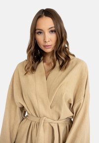 Beżowy szlafrok o strukturze z szerokimi rękawami, w fasonie zapinanym na krzyż, z przewiązywanym paskiem. Gładki materiał, minimalistyczny styl i brak widocznych wzorów lub elementów metalowych.