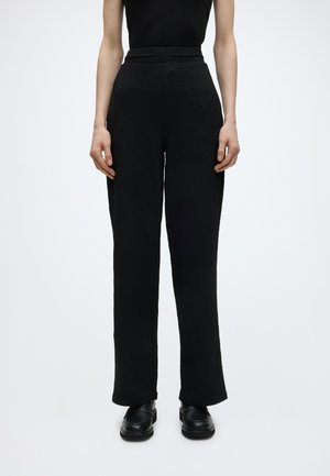 Kvinde iført højttaljede, sorte teksturerede bukser og sorte loafers, der står mod en ensfarvet hvid baggrund.