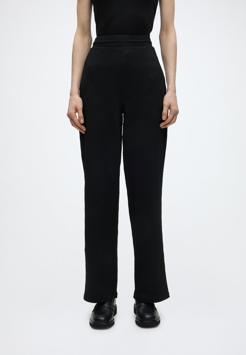 Γυναίκα με μαύρο υφασμάτινο παντελόνι με ψηλή μέση και μαύρα μοκασίνια, που στέκεται μπροστά σε ένα μονόχρωμο λευκό φόντο.