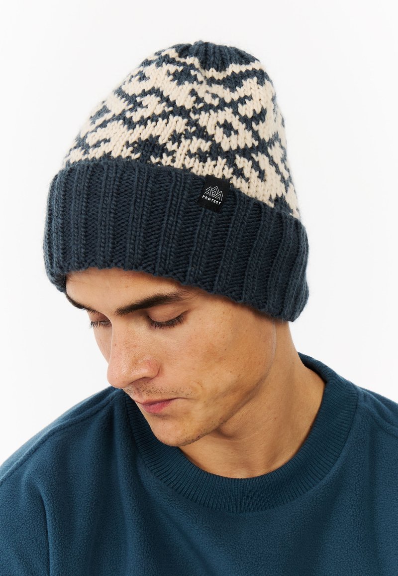 Knit beanie in navy with cream patterned accents, featuring a ribbed cuff and a branded tag on the side. Soft texture and snug fit.