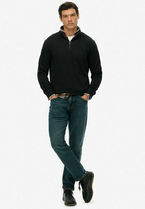 Zwarte zip-nekgolf, donkerblauwe jeans en zwarte schoenen. De pullover heeft een subtiele kraag en de jeans hebben een recht gesneden pasvorm.