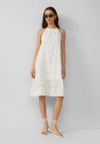 Weißes ärmelloses Kleid mit Lochstickerei, mehrlagigem Saum und gerafftem Ausschnitt. Kombiniert mit weißen Sandalen mit Riemchen und dunklen Sonnenbrillen.