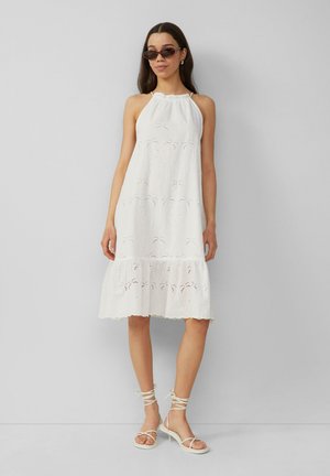 Weißes ärmelloses Kleid mit Lochstickerei, mehrlagigem Saum und gerafftem Ausschnitt. Kombiniert mit weißen Sandalen mit Riemchen und dunklen Sonnenbrillen.