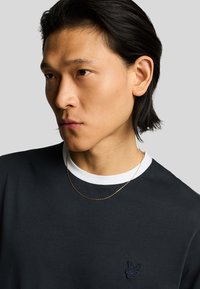 T-shirt in cotone nero con colletto a girocollo bianco, caratterizzato da un piccolo logo ricamato sul petto e una texture liscia. Collana con catena dorata visibile.