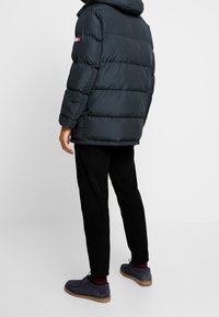 Námořnická péřová bunda s kapucí, s horizontálním prošitím a malou logovou nášivkou na rukávu. Nasučena s černými manšestrovými kalhotami a modrými botami.