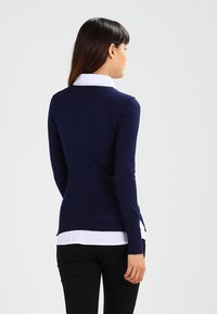 Navy blue pullover sweater with white collar, ribbed hem, and long sleeves, worn over a white shirt, viewed from the back.