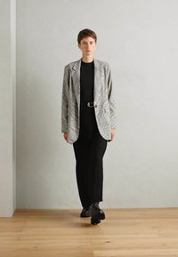 Grijze geblokte blazer over een zwarte jurk, zwarte schoenen met gestructureerde zool, staande op een houten vloer tegen een lichtgrijze muur.