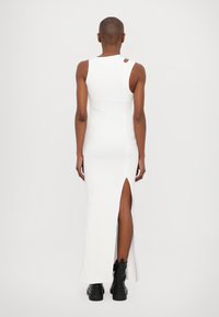 White ribbed maxi dress with a side slit, showcasing a fitted design and a circular cutout at the shoulder. Worn with black ankle boots.