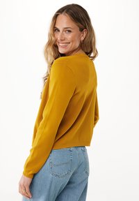 Usmívající se žena s dlouhými vlnitými vlasy, oblečená v hořčicově žlutém svetru a světlých modrých džínách, dívající se přes rameno na bílém pozadí.