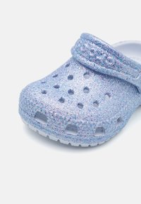 Błyszczące niebieskie klapy z perforowanym wzorem i regulowanym paskiem. Materiał wydaje się lekki i elastyczny, z teksturowaną, błyszczącą powierzchnią.