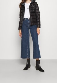 Sort pufferjakke med hætte, hvid t-shirt, vide blå jeans og sorte ankelstøvler med block hæl. Glatte teksturer og lagdelt design.