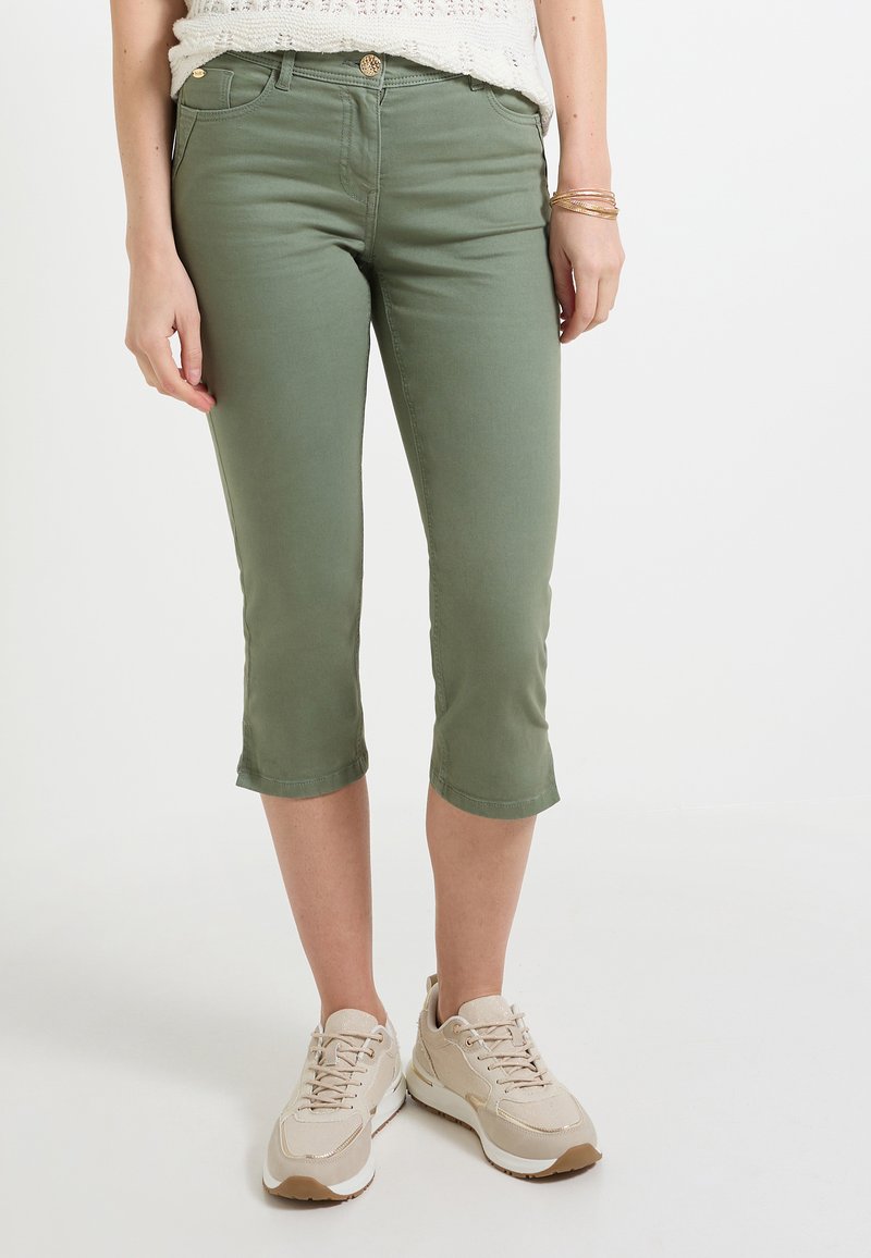 Pantalons capri verts en tissu extensible, avec une coupe slim, présentant un design classique à cinq poches et des ourlets roulés. Portés avec des baskets beiges.