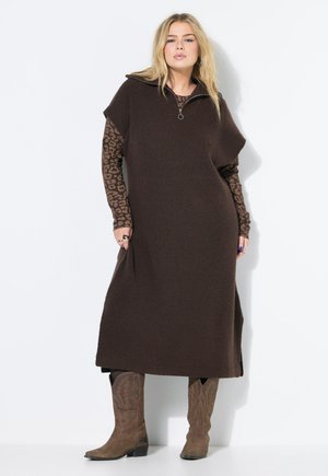 Vestido de punto marrón con un cuello simulado con cremallera, con mangas cortas y bolsillos laterales, superpuesto a una blusa de leopardo de manga larga. Botas marrones.