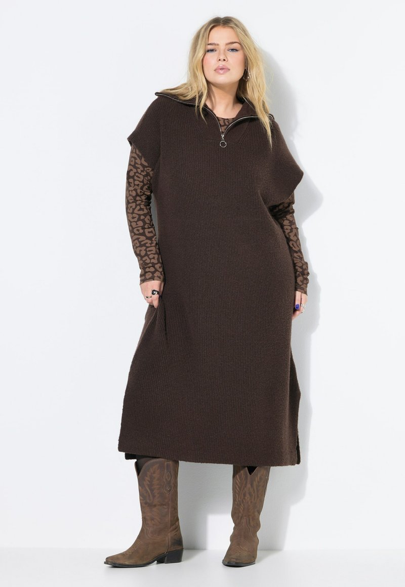 Braunes Strickkleid mit einem Reißverschlusskragen, ausgestattet mit kurzen Ärmeln und seitlichen Taschen, darüber getragen ein langärmeliges Oberteil im Leopardenmuster. Braune Stiefel.