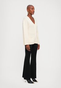 Kobieta stojąca w białym swetrze zapinanym na guziki, czarnych spódnicach dzwonowatych, czarnych czółenkach na obcasie oraz złotych kolczykach w kształcie kół na tle białego tła.