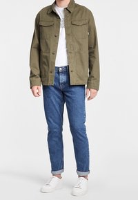 Olive green cotton jacket with chest pockets, paired with blue denim jeans and white sneakers. The jacket features a button closure and collar.