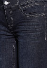 Ciemnoniebieskie dżinsy o dopasowanym kroju. Wśród cech znajdują się kieszenie z nitami, kontrastowe przeszycia oraz klasyczne zapięcie na guzik.