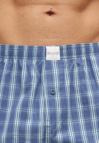 Мъжки торс, носещ сини кареени боксерки Hollister, показващ пъпа с пробито сребърно пръстенче.
