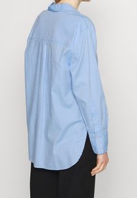 Personne portant une chemise boutonnée à manches longues de couleur bleu clair et un pantalon noir, vue de dos sur un fond clair uni.