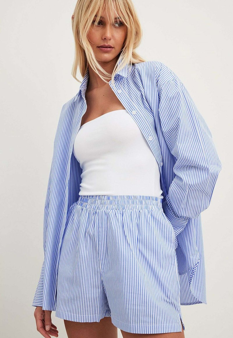 Camicia over-size a righe blu e bianche con maniche lunghe, abbinata a pantaloni shorts a righe coordinate con vita elasticizzata.