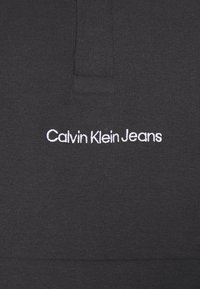 Black fabric close-up with white embroidered "Calvin Klein Jeans" logo centered below stitched seam.