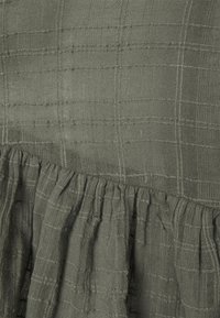 Green textured fabric with a grid pattern, featuring a ruffled hem. Material appears lightweight and breathable, suitable for casual wear.