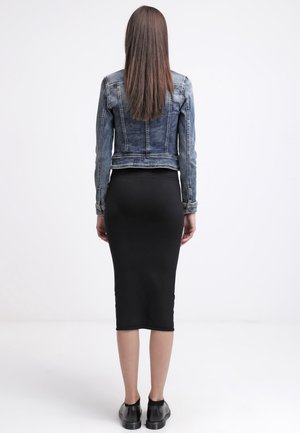 Kobieta z długimi prostymi włosami ubrana w skróconą kurtkę dżinsową, czarną ołówkową spódnicę midi i czarne buty, stojąca na białym tle.