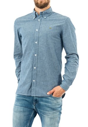 Uomo che indossa una camicia blu a maniche lunghe abbottonata con un piccolo logo verde di coccodrillo, abbinata a jeans azzurri, mano in tasca.