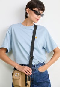 T-shirt en coton bleu clair avec un petit logo orange, associé à un short en denim bleu foncé et un sac bandoulière beige avec des accents noirs.