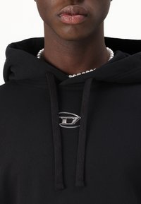 Haut à capuche noir avec un accent en métal argenté à l'avant. Comprend des cordons de serrage, des poignets côtelés et une texture douce en coton. Vue en gros plan.