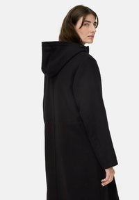 Schwarzer Kapuzenmantel aus strukturiertem Stoff, mit lässiger Passform, versteckten Seitentaschen und einem Knopfverschluss am Hals.