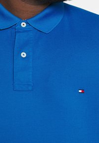 Blå bomullspolo med en strukturerad tyg. Har en tvåknapps placket och ett broderat logo i röd, vit och svart på bröstet.