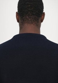 Marineblauwe gebreide trui met een ronde halslijn, voorzien van een geribbelde textuur en een gladde afwerking bij de kraag en de zoom. Achteraanzicht weergegeven.