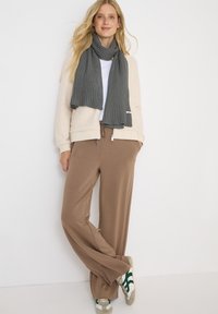 Beige fleecejakke med ribbet grå tørklæde, hvid t-shirt og løse brune bukser. Grå og grønne sneakers med en struktureret finish.
