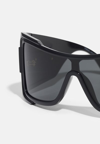 Sorte overdimensionerede solbriller med en geometrisk ramme, mørke linser og en blank plastikfinish. Bemærkelsesværdige detaljer inkluderer udtalt kant og et robust design.