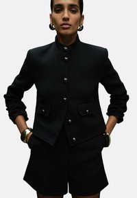 Black wool blazer with a high collar, front buttons, and side pockets; paired with tailored black shorts and a metallic bangle on the wrist.