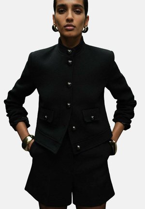 Black wool blazer with a high collar, front buttons, and side pockets; paired with tailored black shorts and a metallic bangle on the wrist.