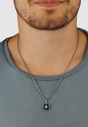 Silver necklace with a circular compass pendant featuring N, S, E, W directions, worn over a gray T-shirt with a crew neckline.
