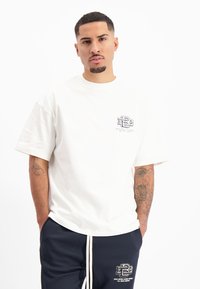 Witte T-shirt met korte mouwen en een ronde hals, met een klein zwart logo op de linkerkant van de borst. Draagt het met marineblauwe trainingsbroek met een logo.