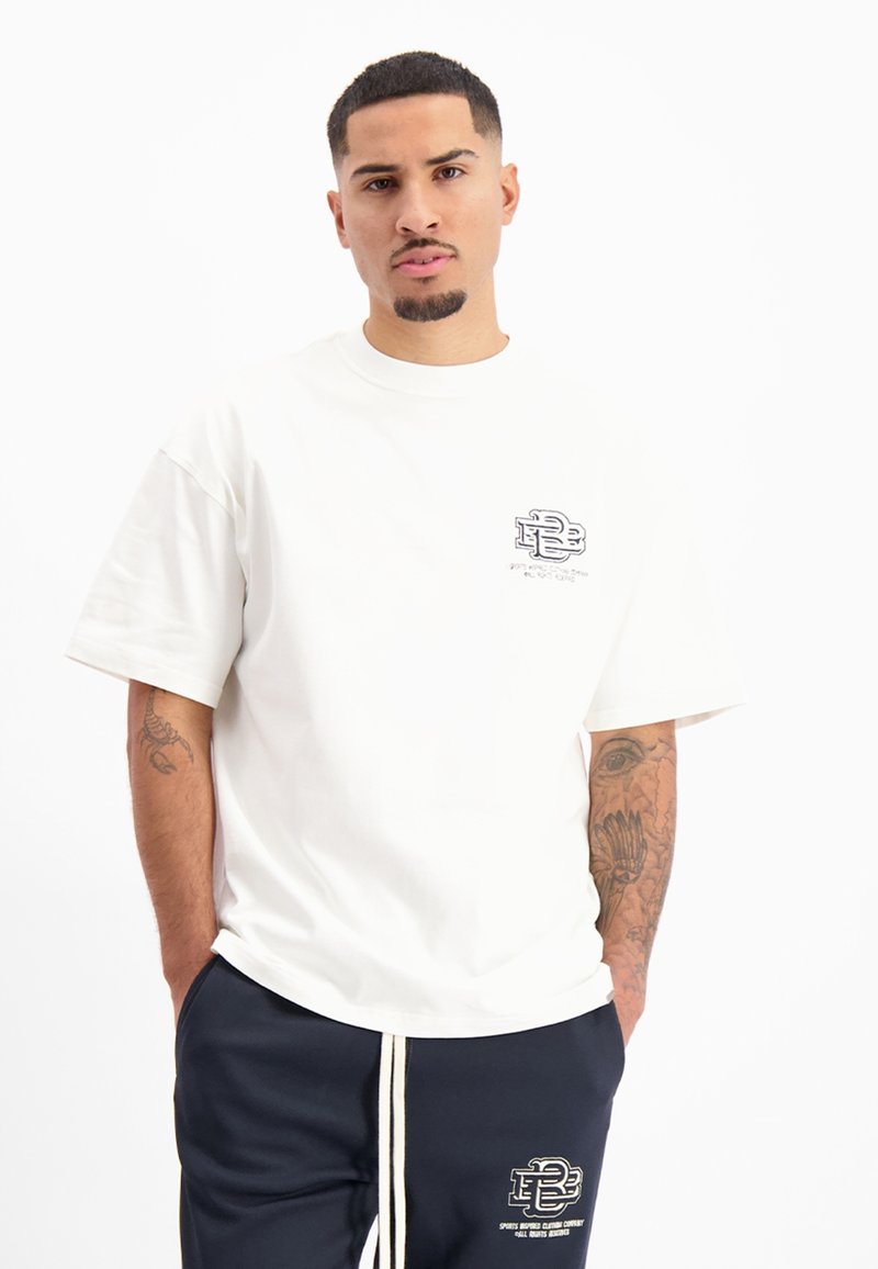 Witte T-shirt met korte mouwen en een ronde hals, met een klein zwart logo op de linkerkant van de borst. Draagt het met marineblauwe trainingsbroek met een logo.