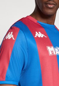 Μπλε και κόκκινη αθλητική φανέλα με κοντά μανίκια, στρογγυλή λαιμόκοψη, υφασμάτινη υφή και λευκό λογότυπο στους ώμους και στο μπροστινό μέρος.