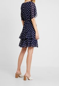 Marinblå polkadotklänning med genomskinlig, svävande tyg, volang nederkant och bälte i midjan. Kombinerad med vita vävda mule-skor och blockklackar.